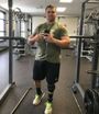 Man with a prosthetic leg giving a thumbs up in a gym. He is wearing workout clothes and holding a phone.