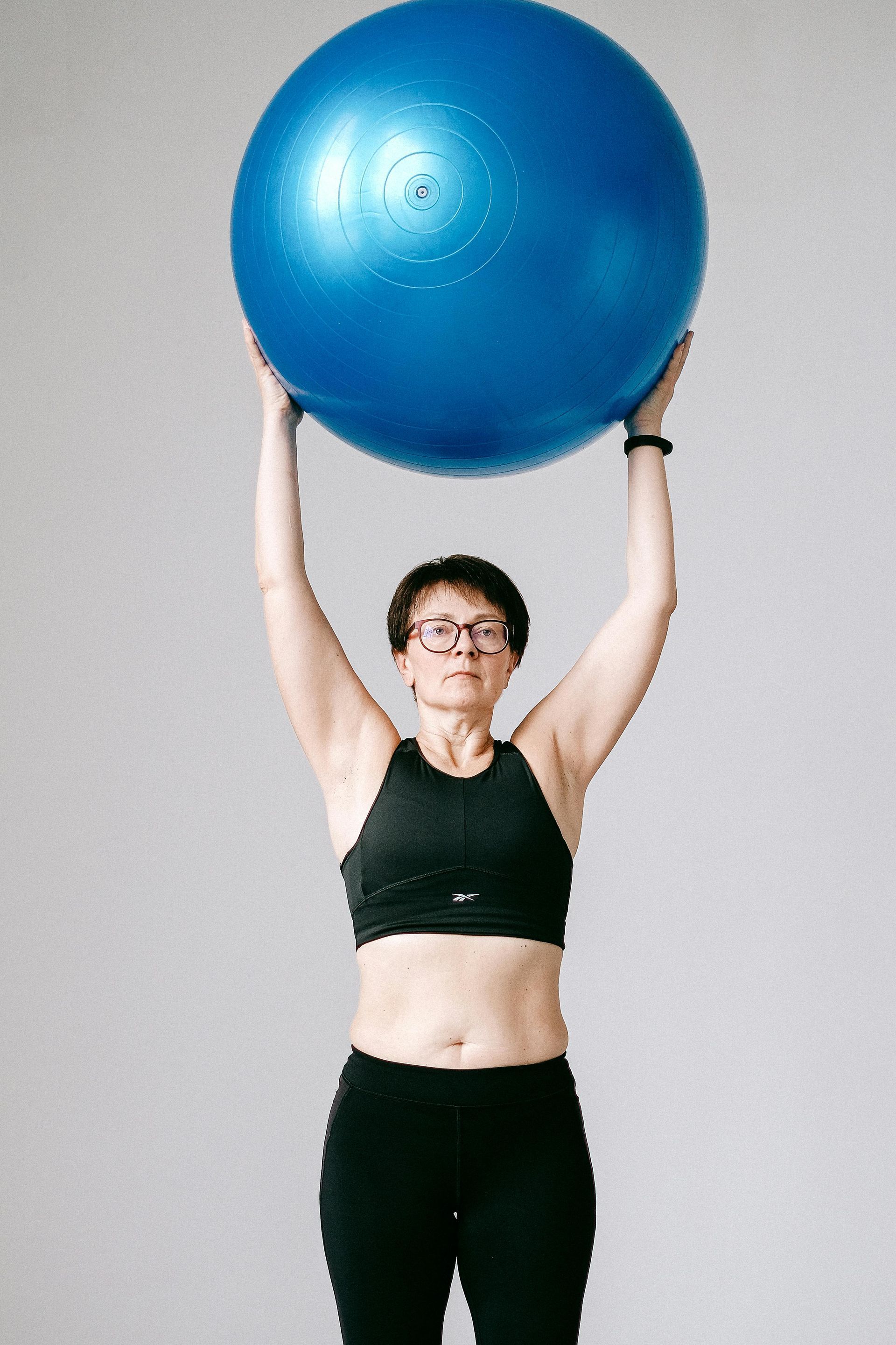 Woman in athletic wear holding blue exercise ball overhead.