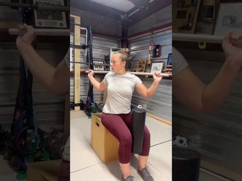 Woman exercising with a barbell, seated on a box, using a foam roller. Inside a garage-like space.