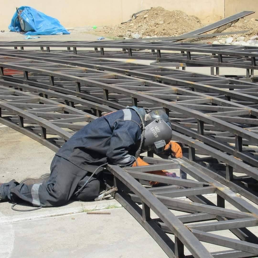 Un soldador con mono azul hace saltar chispas de metal en un taller con poca luz, enmarcado por vigas de acero y maquinaria.