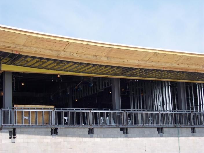 A construction site showing a steel frame structure with a plywood roof overhang above a metal balcony railing.