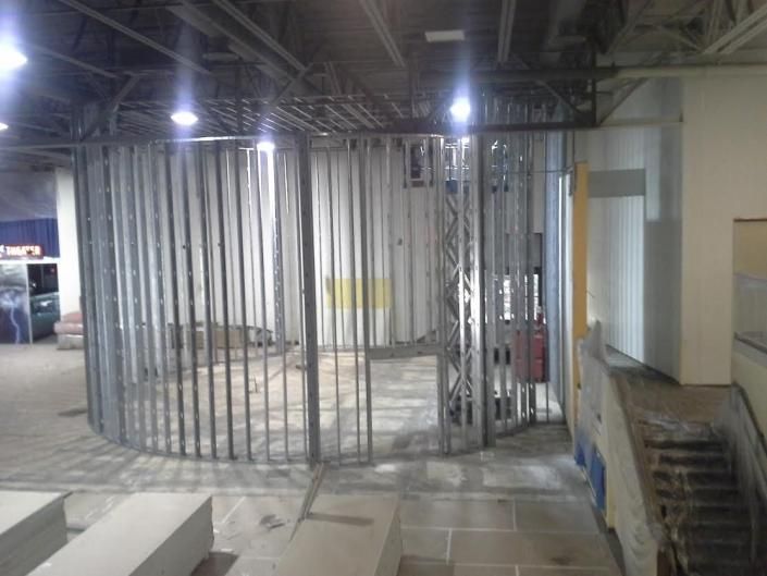 A circular metal stud frame for a wall under construction in a large, unfinished indoor space with concrete flooring.
