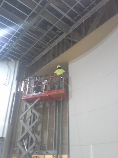 A worker in a high-visibility vest on a red scissor lift, installing materials on a high interior wall.