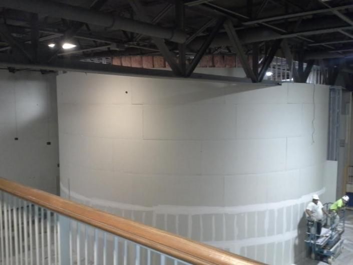 Workers on a scaffold install curved drywall panels on a large interior wall in an industrial construction space.