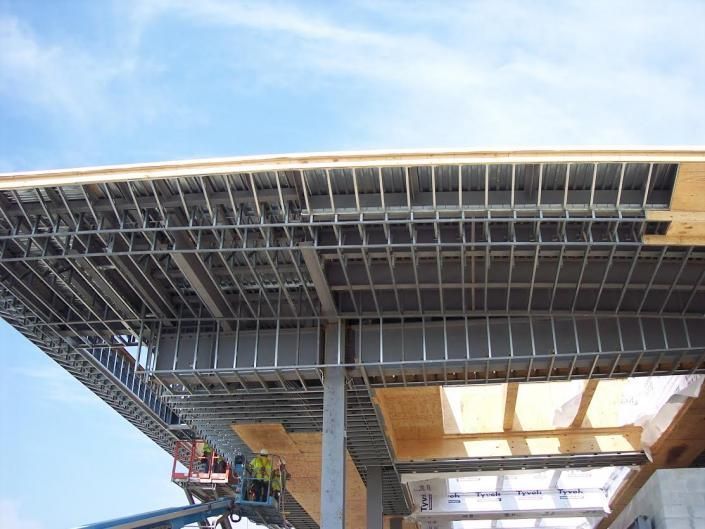Workers in a lift install metal framing for a large building's overhanging roof, revealing wooden beams and steel supports.
