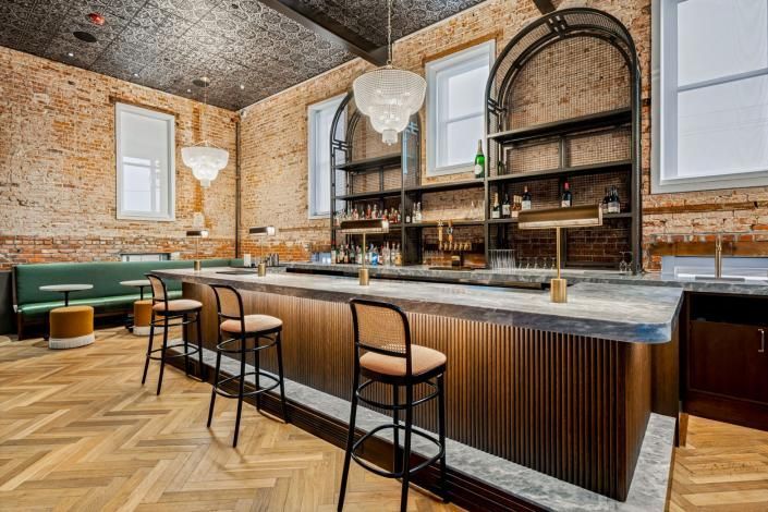 A bar with a stone counter, three stools, and green bench seating in a room with exposed brick walls and a tin ceiling.