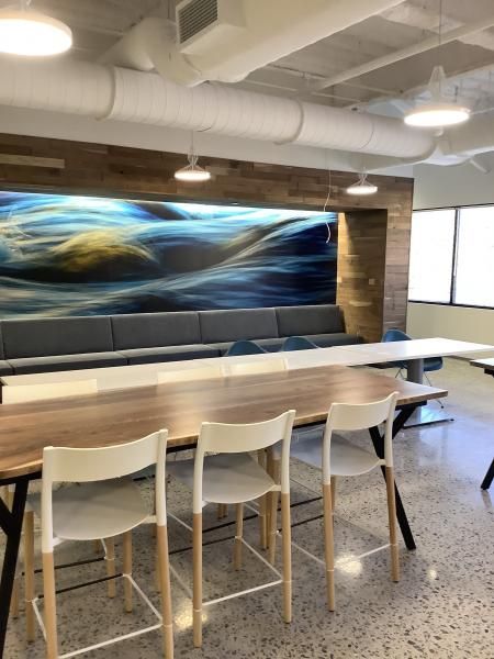 Modern office space with a wood-accent wall, a blue abstract art mural, long tables, and white stools on concrete floors.