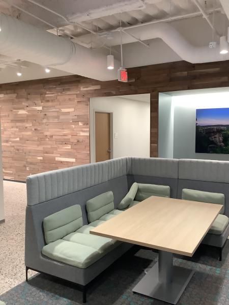 A modern office break area featuring a light wood table with green cushioned booth seating against a wood-paneled wall.