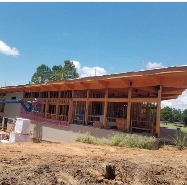 A wooden-framed, partially constructed building stands on a dirt construction site under a bright, clear blue sky.