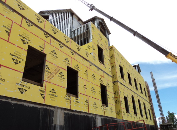 A construction site with a yellow sheathed multi-story building frame and a crane arm overhead against a blue sky.