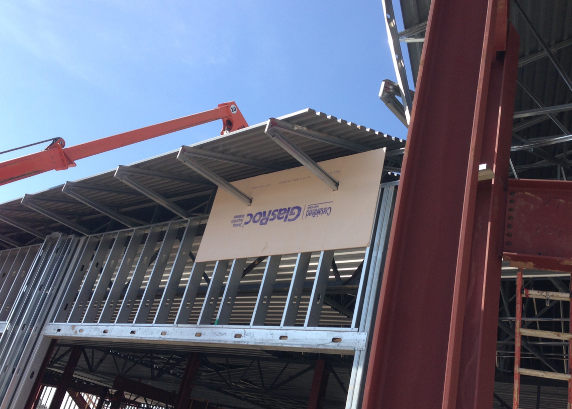 Construction site featuring a metal frame structure, a Glasroc brand panel, and a red crane arm against a blue sky.