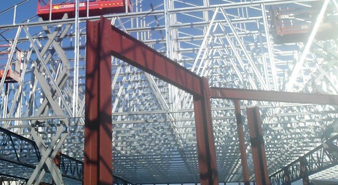 Red structural steel beams frame a construction site with extensive metal roof trusses and elevated work platforms.