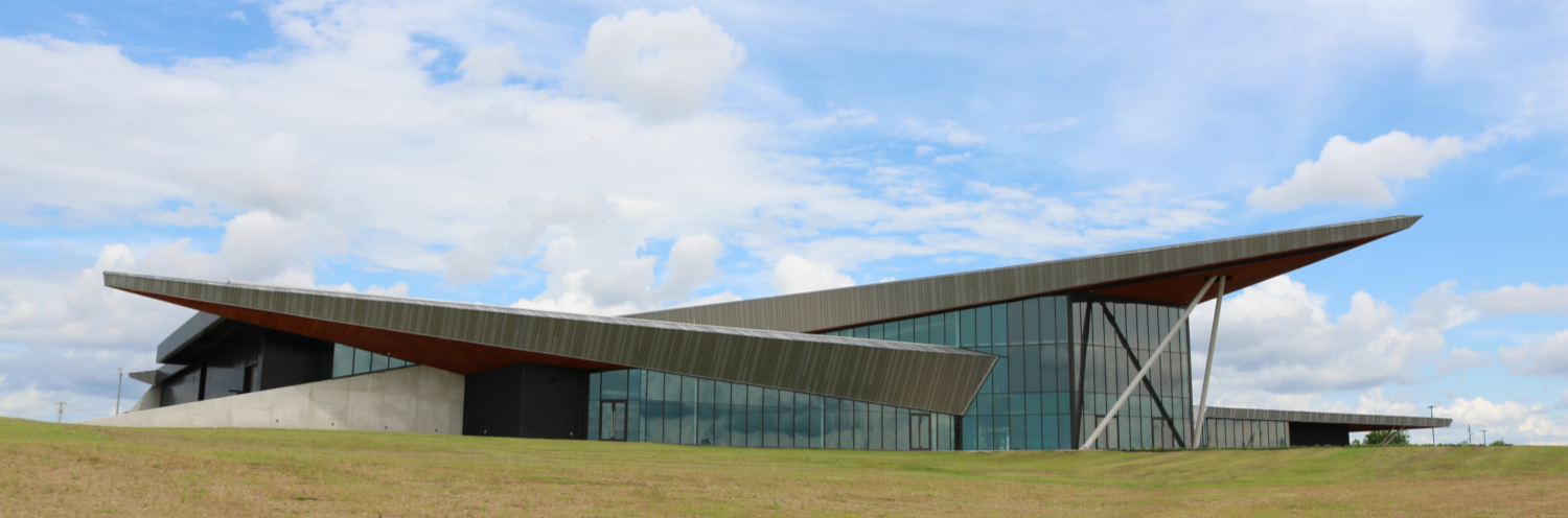 Modern building with a large, slanted dark roof and expansive glass walls, set in a grassy field under a cloudy sky.
