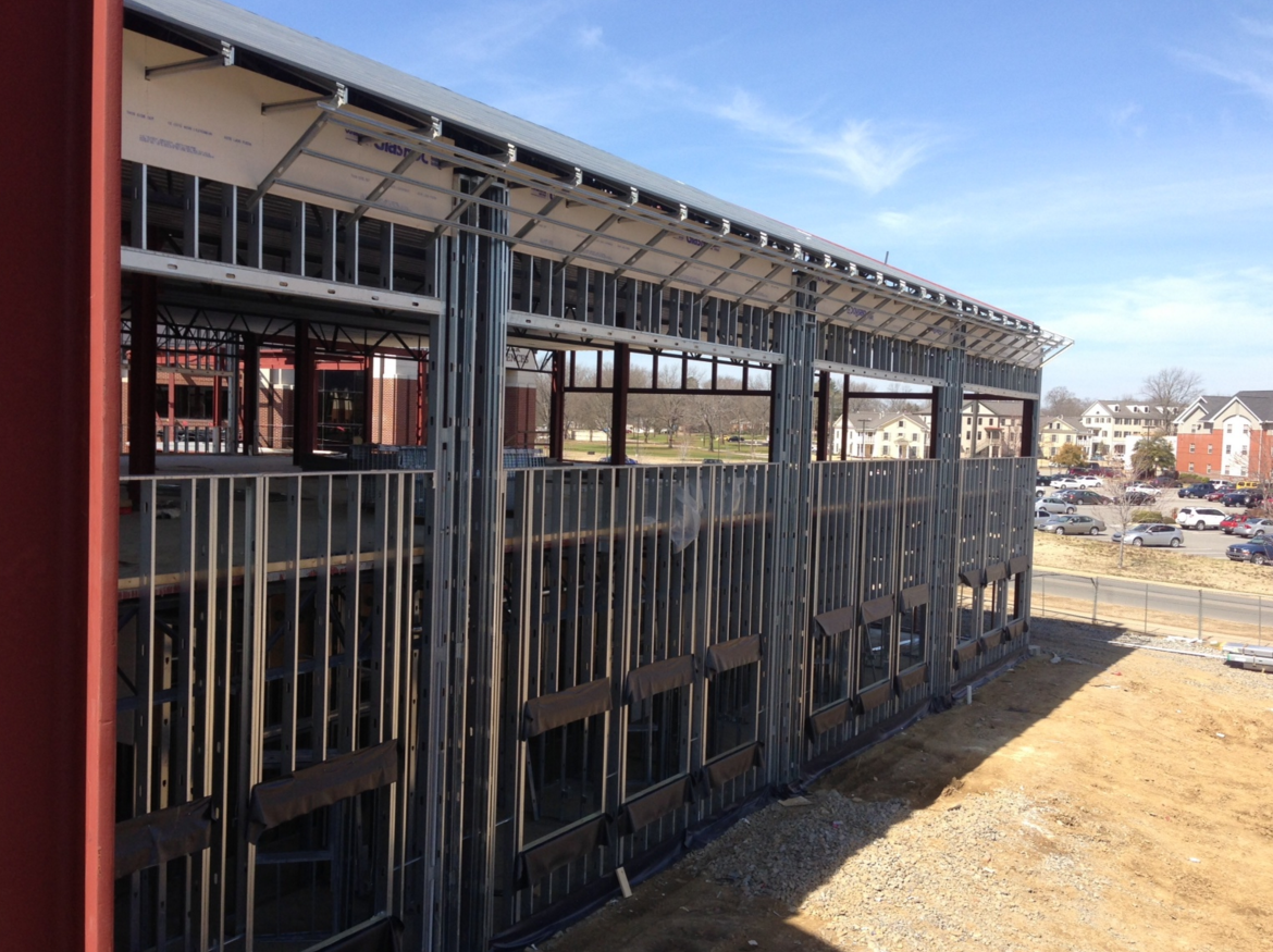 The steel frame of a building under construction, showing vertical studs and exterior siding against a clear blue sky.