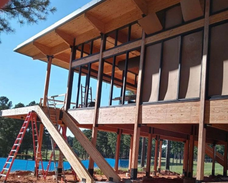 A modern timber-frame house under construction on stilts, featuring large glass windows and an exterior wooden staircase.