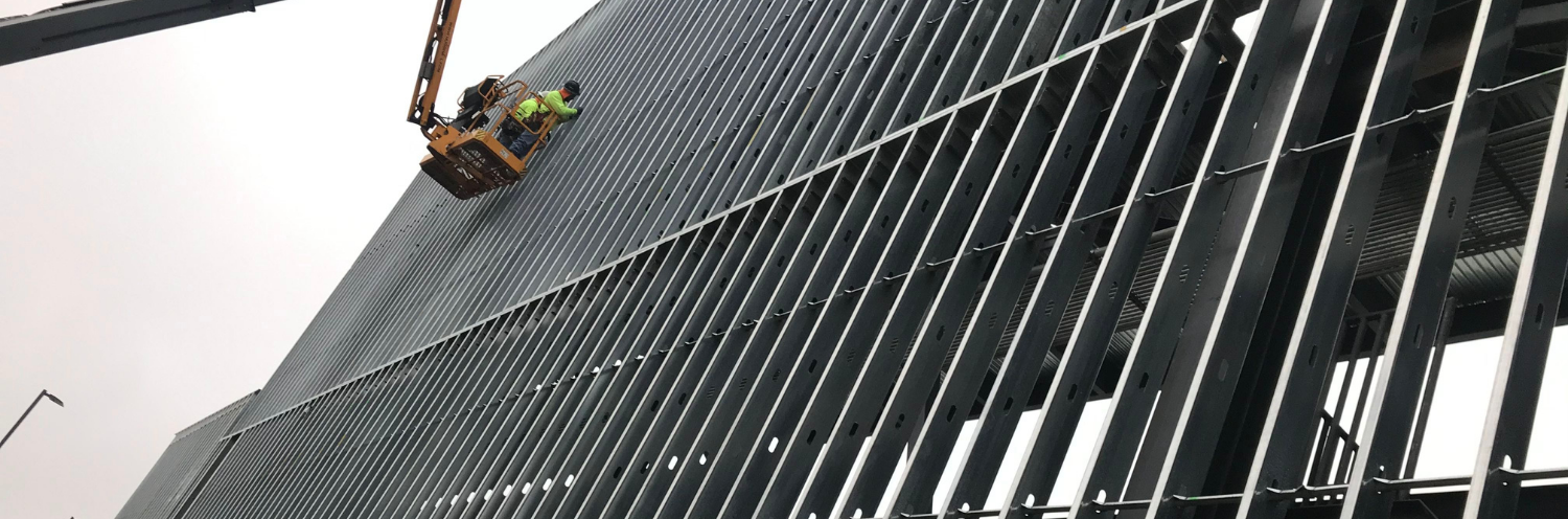 A worker in a lift basket installs gray metal panels on the exterior of a building under construction.