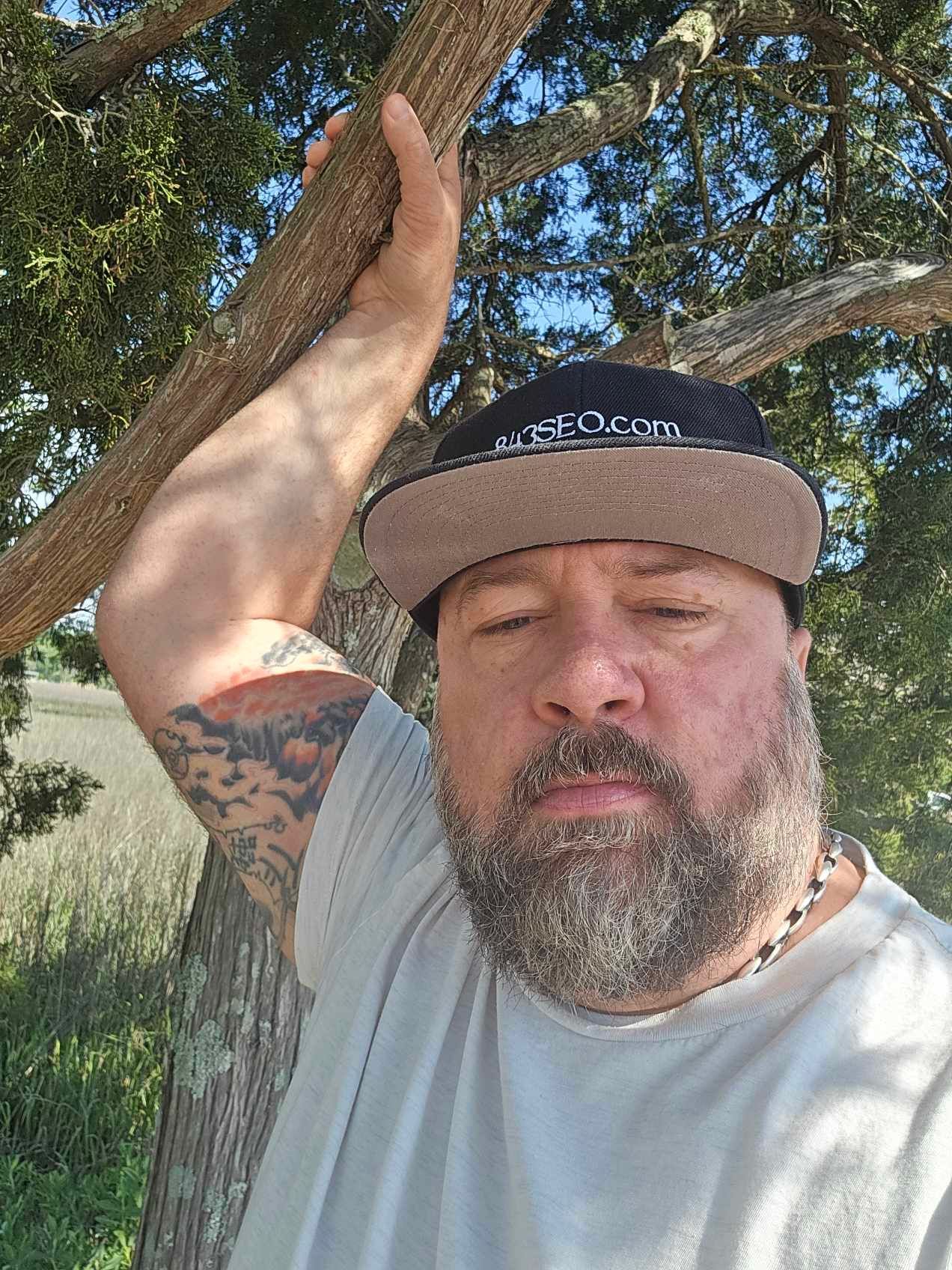 Man with a beard and cap, leaning against a tree branch outdoors. Tattoo on arm.