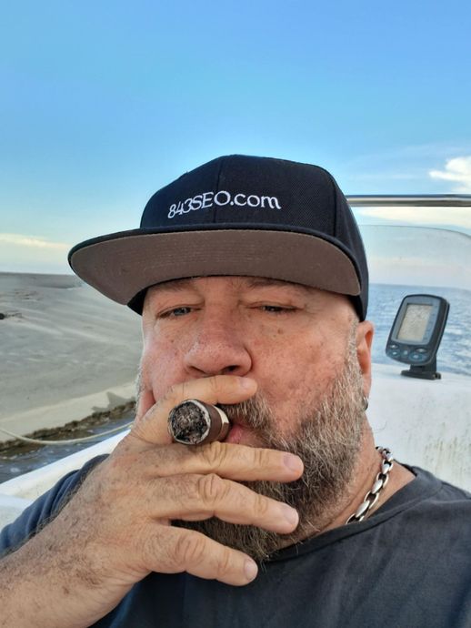 Man smoking cigar on a boat, wearing hat, blue sky background.