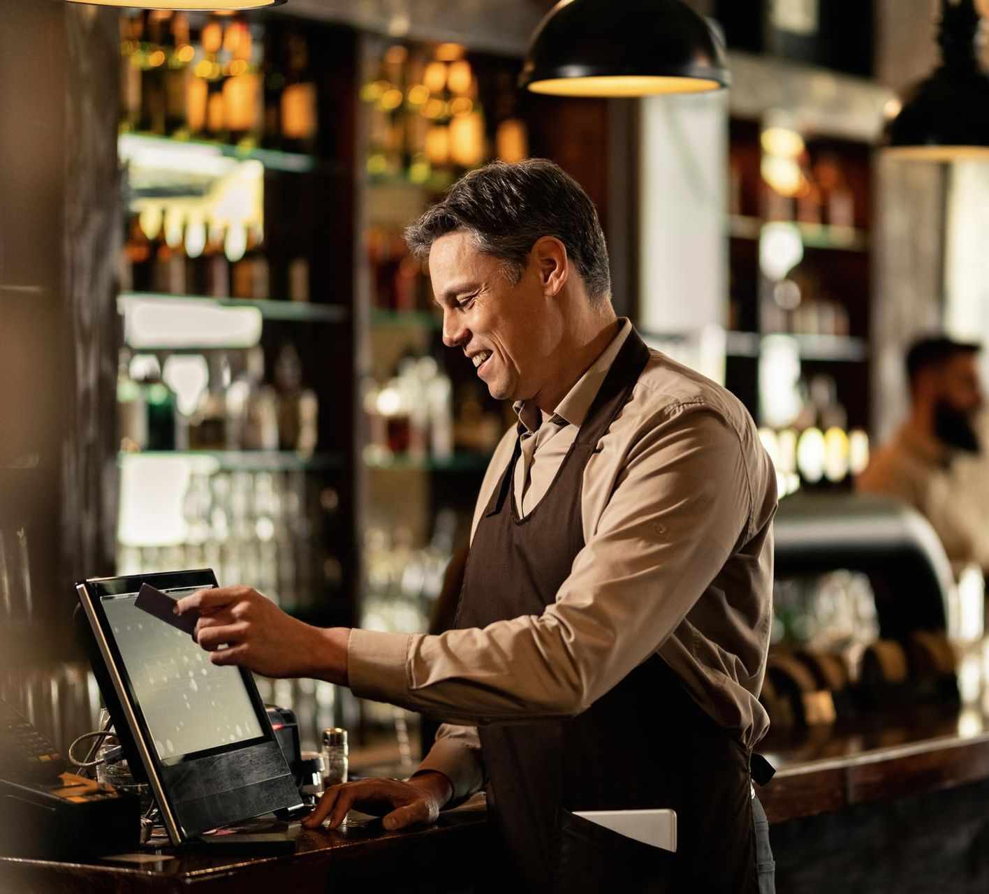 A smiling bartender in a brown apron uses a point-of-sale system at a bar, lit by overhead lights.