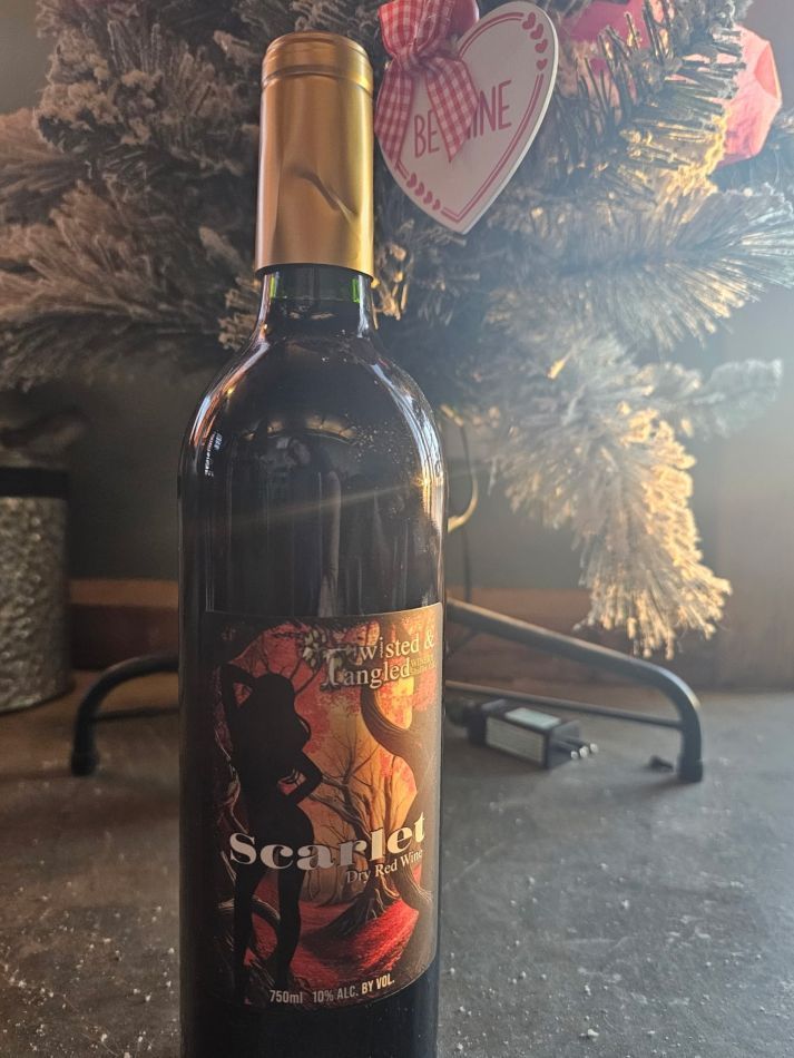 A bottle of Scarlet Letter wine stands on a dark surface in front of a decorated Christmas tree with a heart ornament.