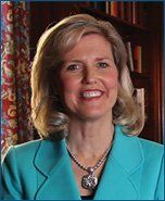 A woman in a blue jacket and necklace is smiling in front of a bookshelf.