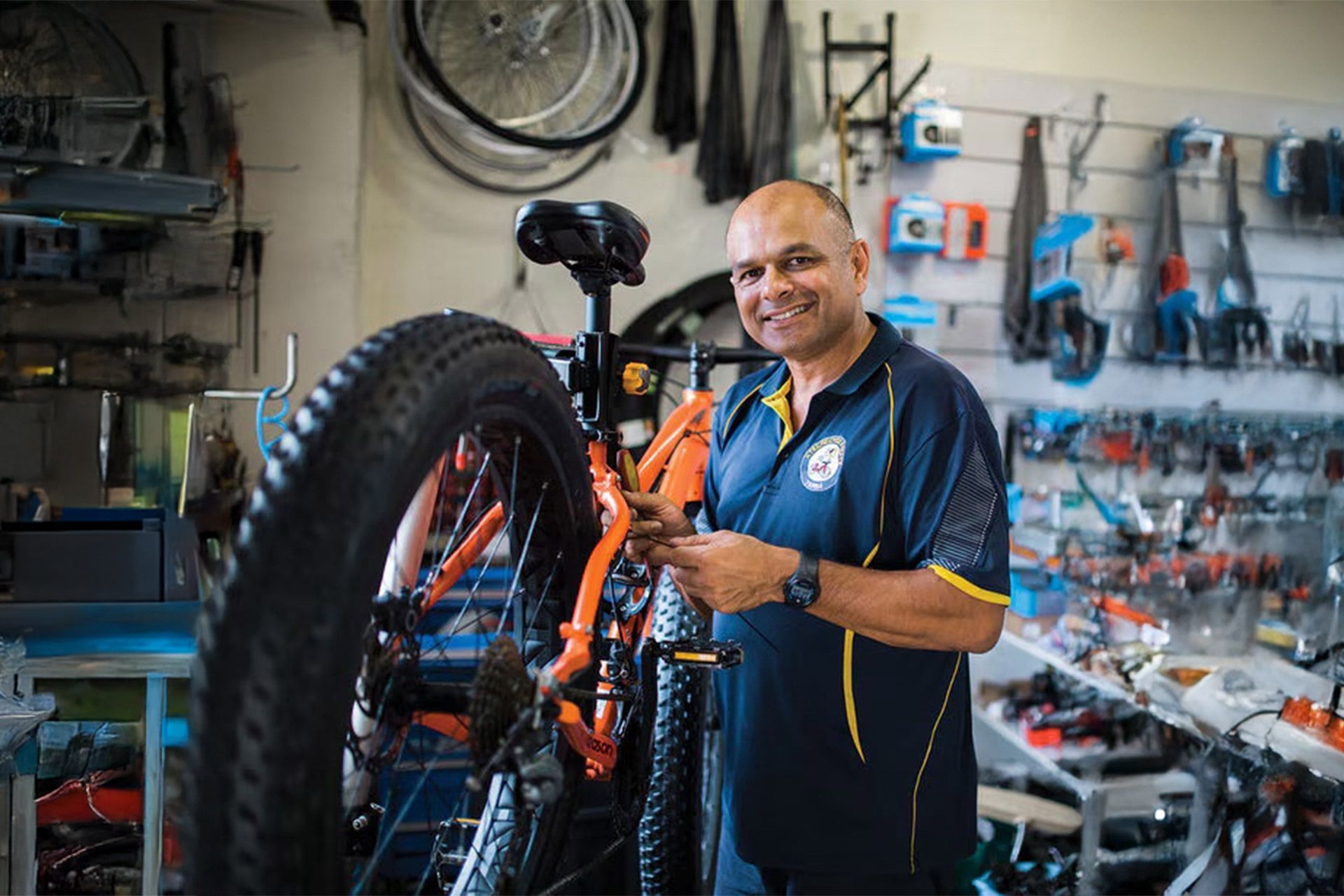 Man fixing a bicycle tire in a bike shop; he smiles, wearing a blue and yellow shirt.