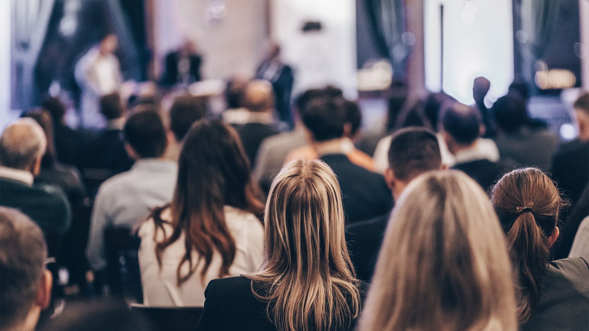 Audience seated in a dim conference room, facing a stage with speakers.