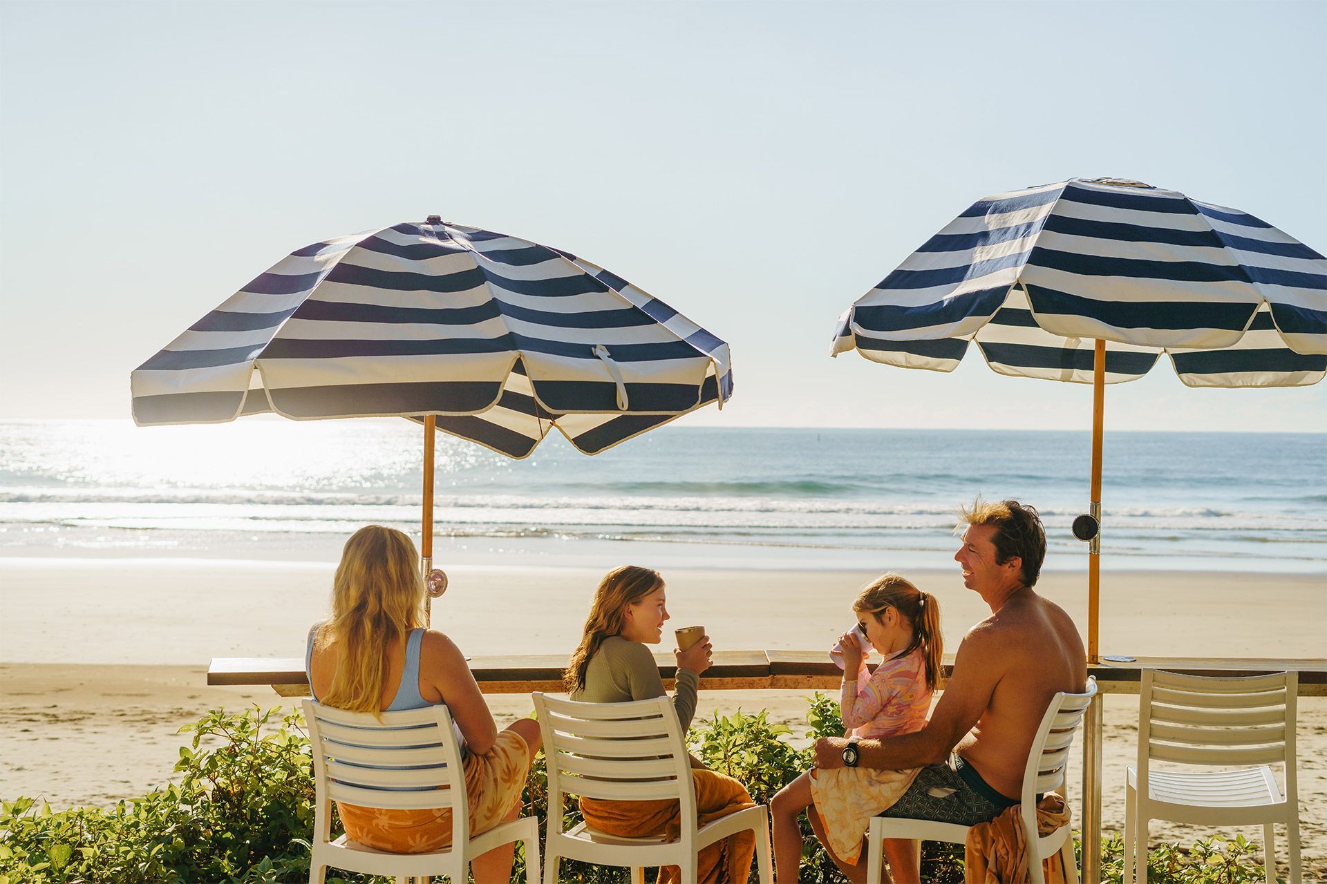 Family under striped umbrellas at a beachside table, enjoying drinks with the ocean in the background.