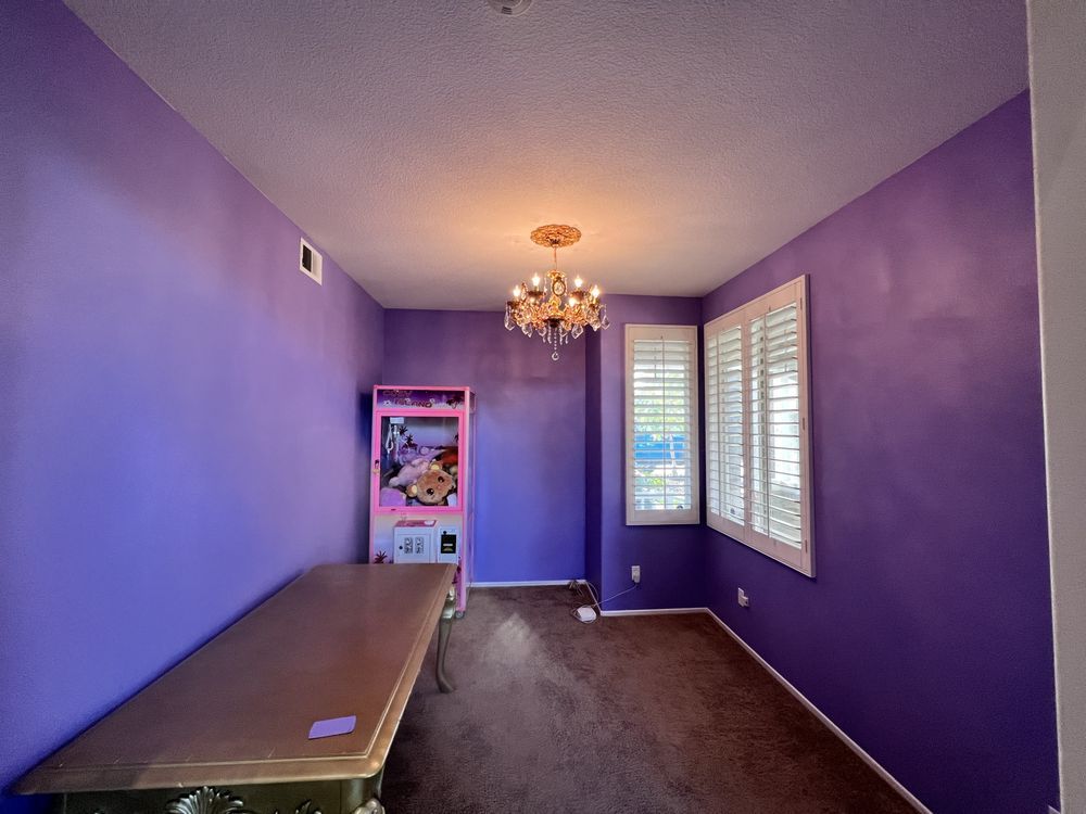 Purple-walled room with brown carpet, chandelier, arcade game, table, and window with shutters.