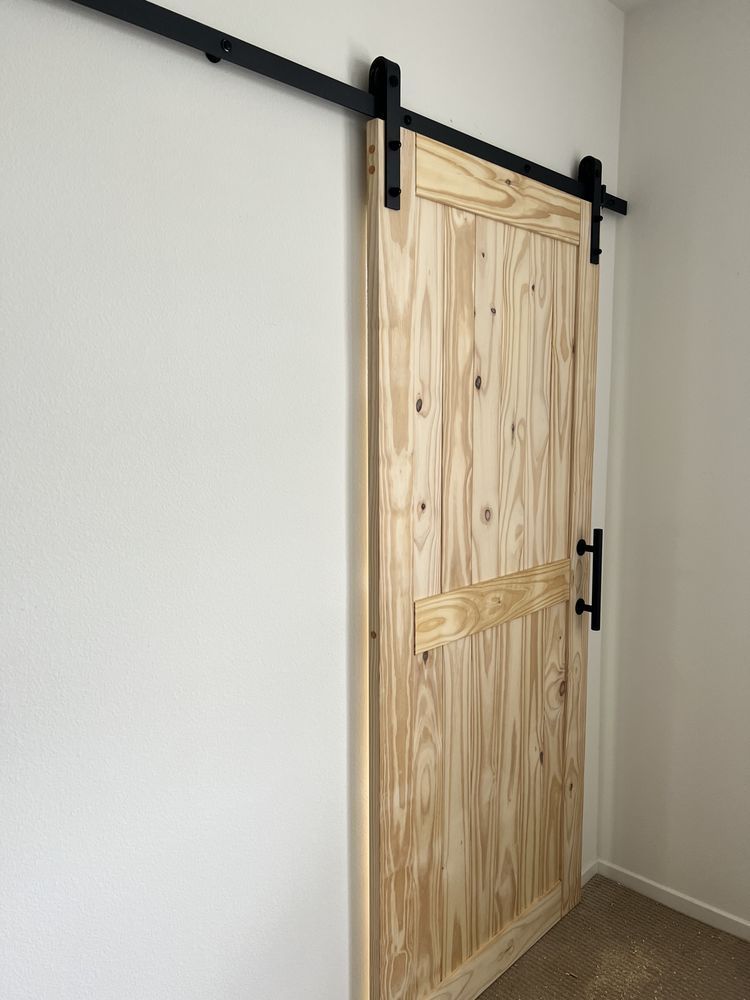 A light wood barn door with black hardware hangs on a white wall.