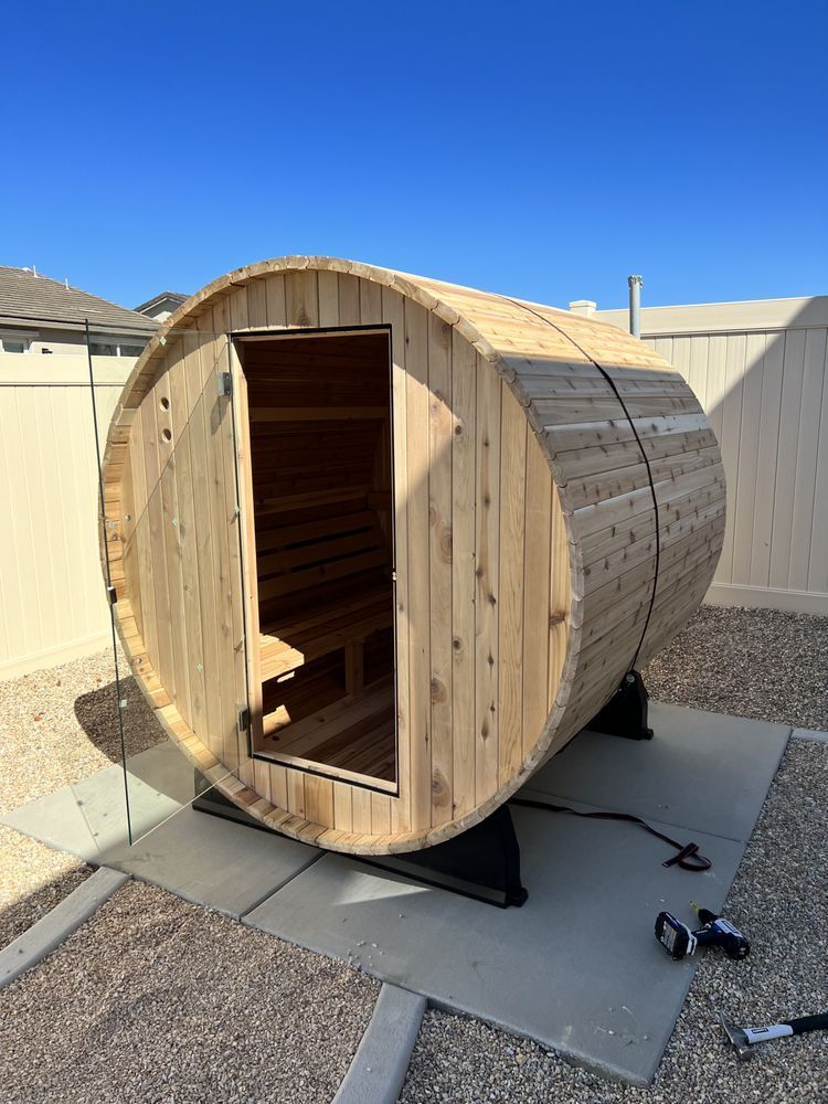 Wooden barrel sauna with open door, outdoors on a concrete pad, against a white fence.