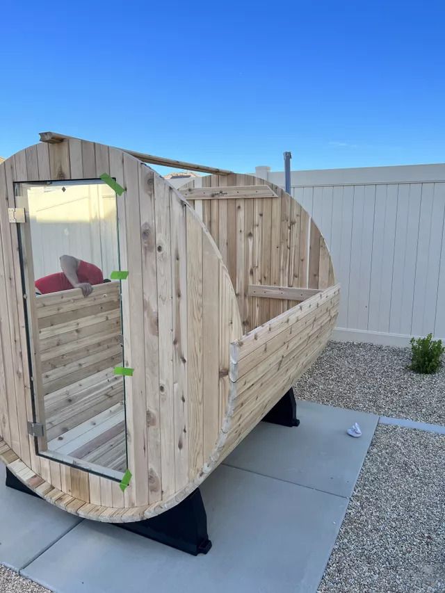 Wooden barrel sauna under construction on a concrete patio, against a white fence and blue sky.