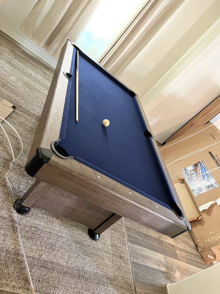 Pool table with a blue felt playing surface, cue stick, and white cue ball. Brown wood-like frame.