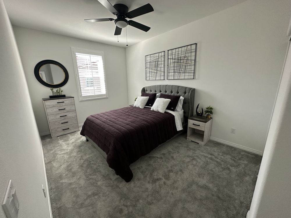 Bedroom with gray carpet, white walls, and a bed with a dark purple quilt and headboard; a chest of drawers, mirror, and two art pieces are visible.