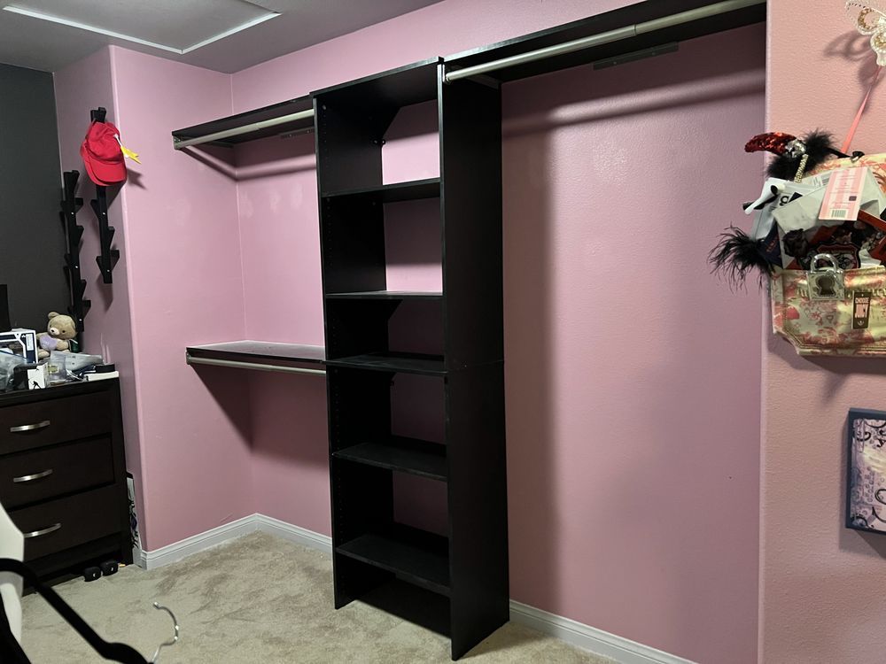 Closet interior with dark shelving, hanging rods, and pink walls. A dark dresser is in the left corner.