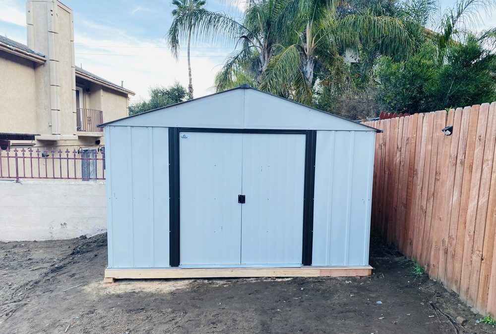 Light blue metal shed with black door trim and a wood base, set between a fence and a building.