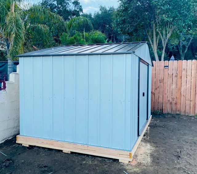 Light blue shed with dark roof and door, set on a wooden base in a backyard.