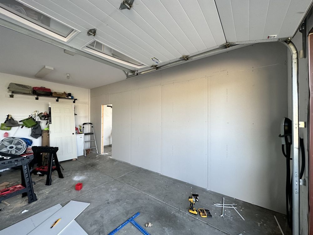 Garage interior with drywall on one wall, open garage door, tools on the floor.