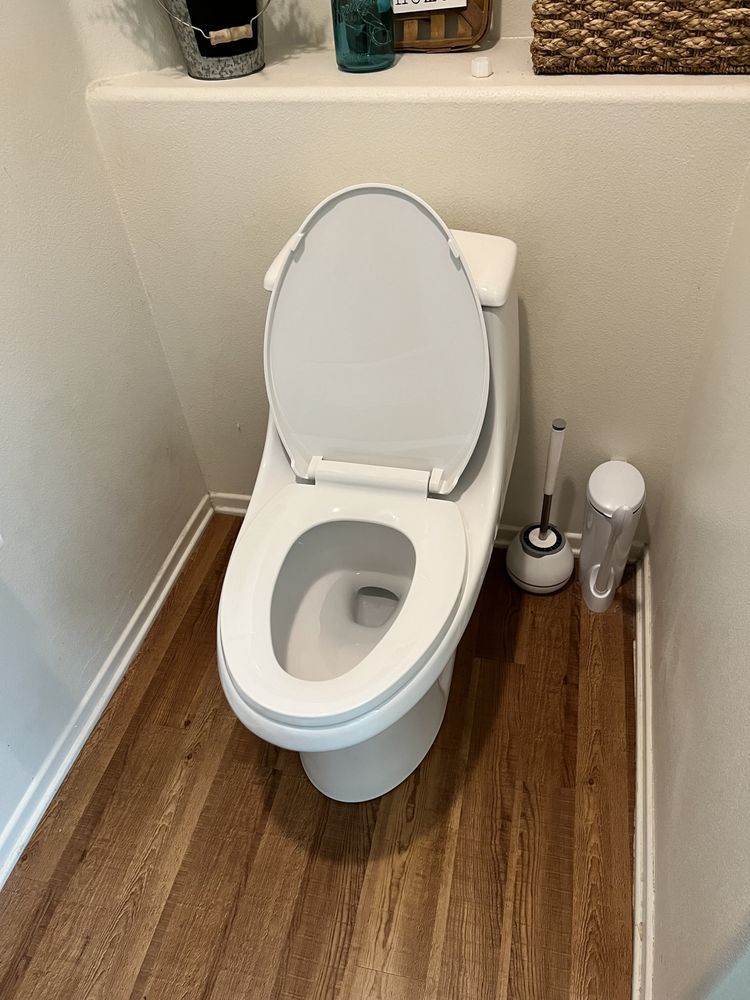 Toilet with the lid up in a small bathroom with wooden flooring. Toilet brush to the right.