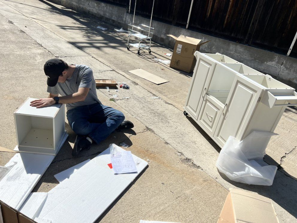 Man kneels assembling a white cabinet outdoors on a paved area. Box and parts surround him.