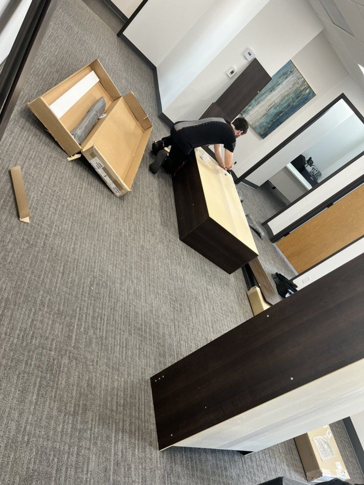 Person assembling a brown desk in an office setting. Parts and open box are on the floor.