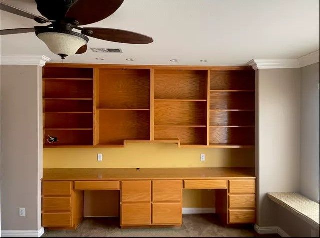Built-in wooden desk and shelving unit. Light brown cabinets and shelves against beige wall, with a ceiling fan.