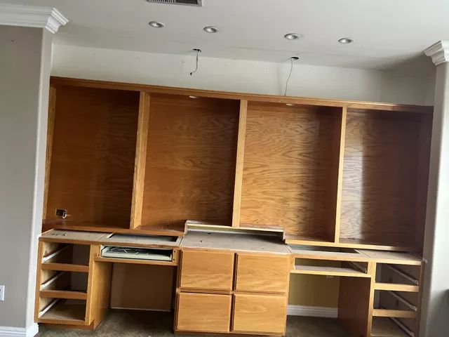 Built-in wooden desk with drawers and upper shelves. Unfinished wood against beige walls, recessed lighting.