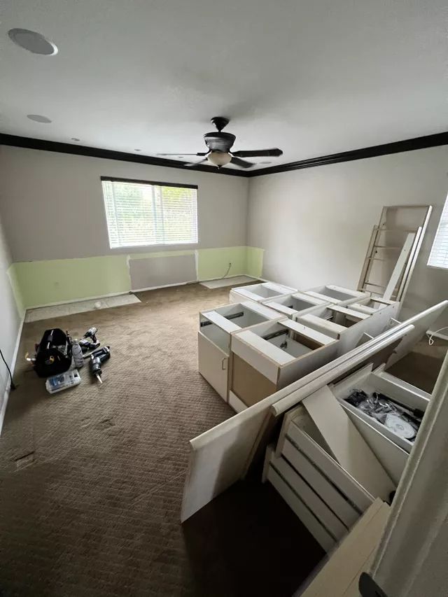 Room under renovation with cabinets and supplies; brown carpet, light green and beige walls, black trim, and ceiling fan.