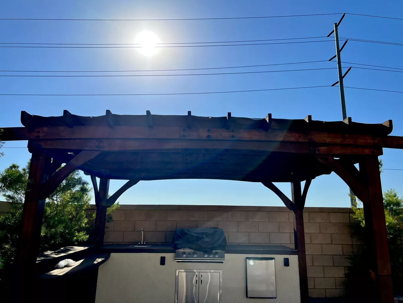 Outdoor kitchen under a wooden pergola on a sunny day. Includes a grill and storage.
