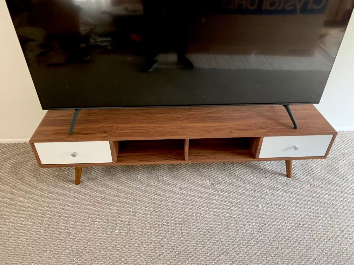 TV on a mid-century modern style wood and white TV stand with two drawers and open shelves on a beige carpet.