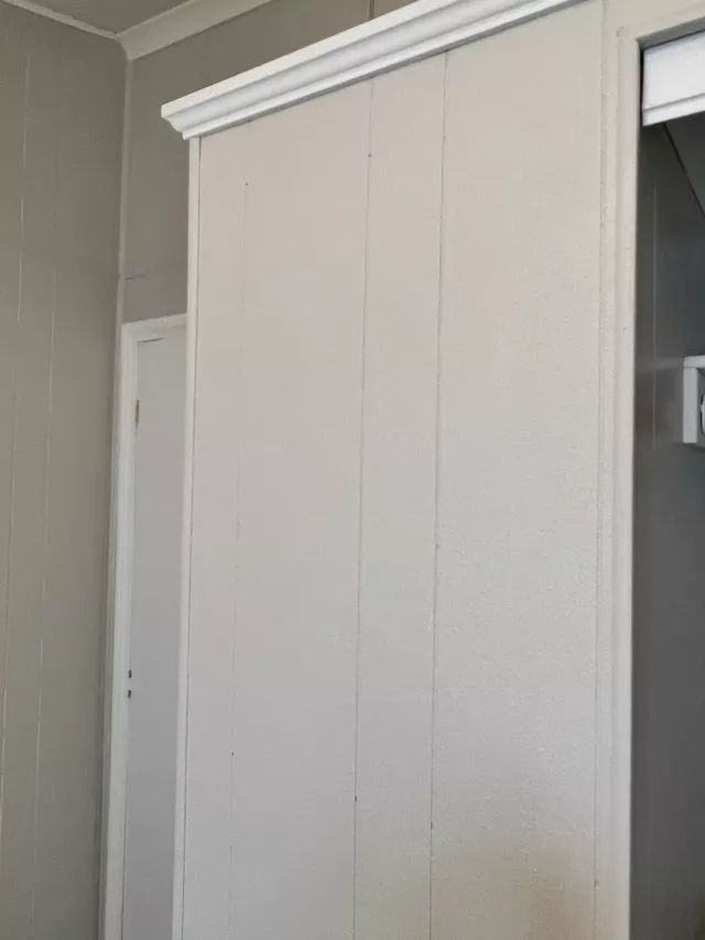 White paneled wall with decorative trim, partially blocking a doorway. Gray walls are visible.