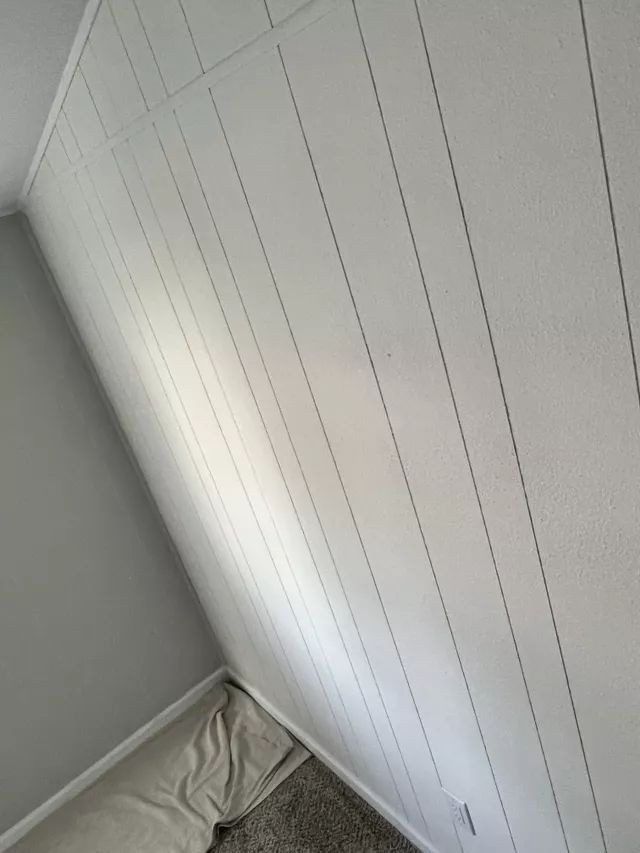 White vertical paneling on a wall in a room. Gray carpet and a beige cushion are visible.