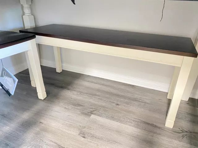 Wooden bench with a dark brown top and cream-colored legs, set against a white wall.