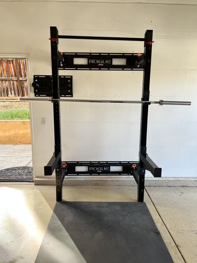 Black Rogue squat rack with barbell in a garage setting, mounted on a wall.